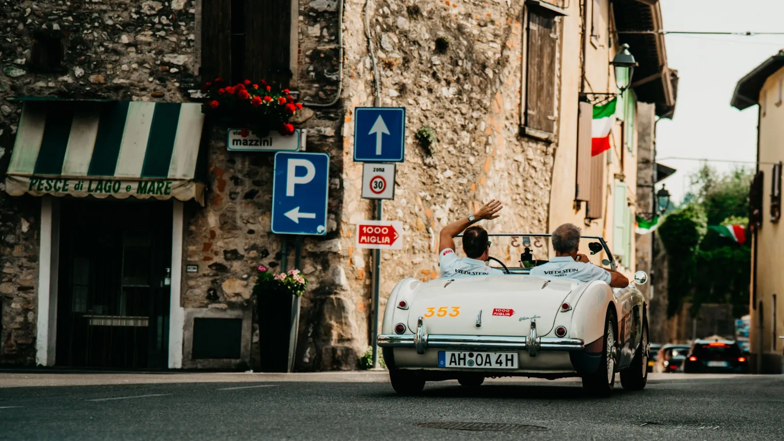 Rear view of a vintage white classic convertible sports car bearing license plate AH 0A 4H driving past rustic stone buildings during the 1000 Miglia rally. (Photo: Apollo Tyres, Vredestein)
