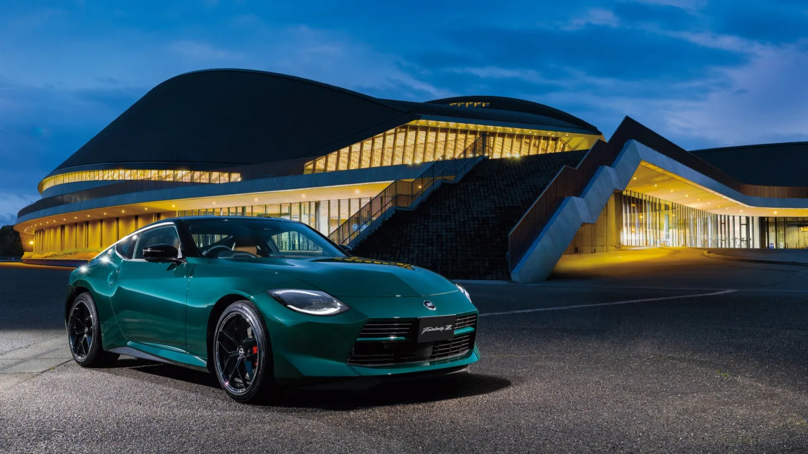 Front three-quarter view of a dark green Nissan Fairlady Z Nismo sports coupe parked on asphalt at night with a large illuminated modern building in the background.