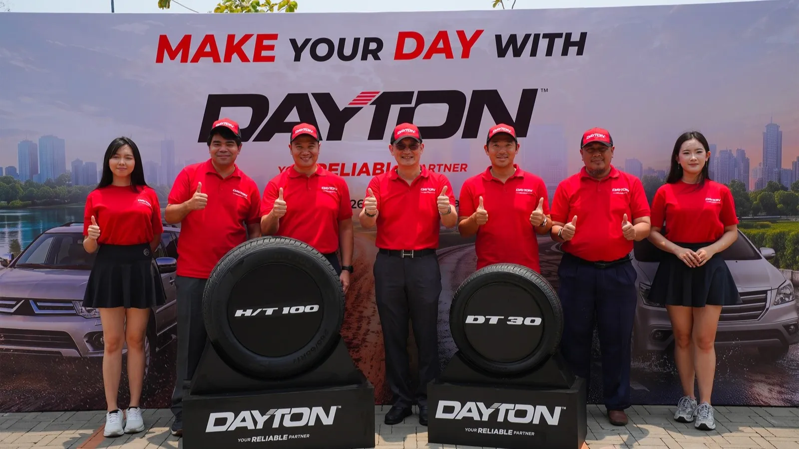 Executives wearing red Dayton caps and shirts giving thumbs-up gestures while standing behind two display tyres, the H/T100 and DT30, in front of a red "Make Your Day With Dayton" backdrop.